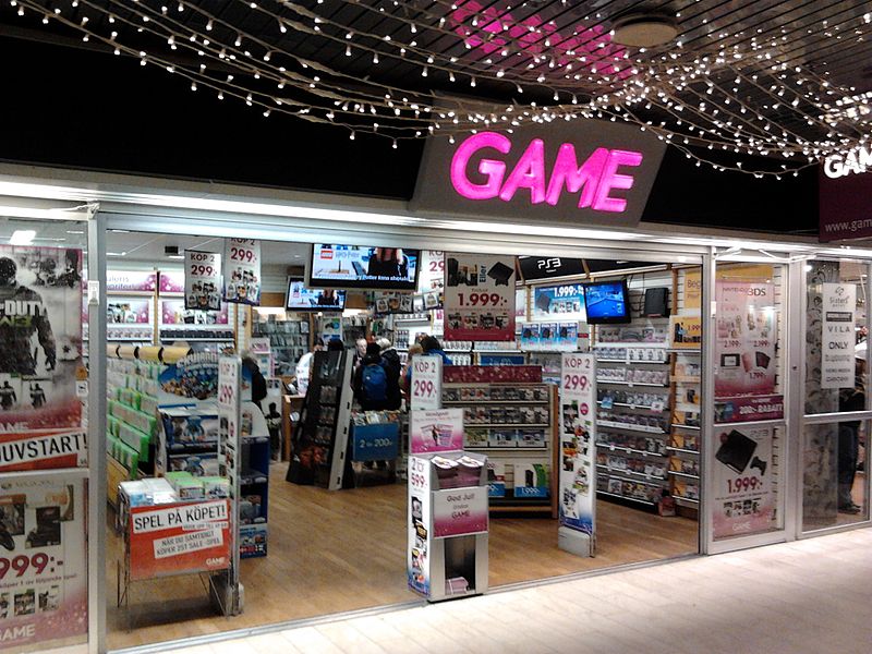 Puerta principal de una tienda de video juegos con diversos estantes, pantallas y carteles con los precios de los artículos.