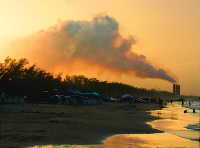 La imagen muestra una playa y al fondo las chimeneas de una fábrica echando humo negro.
