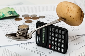 Imagen de una cuchara en equilibrio sobre una calculadora. A un lado tiene clavada una patata, en el otro sostiene monedas.