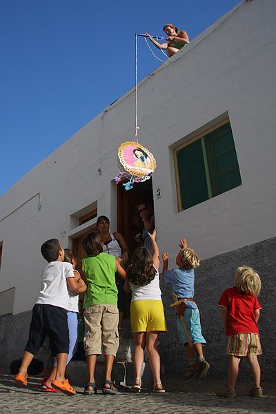 La imagen muestra una señora en la azotea de una casa. Sujeta una piñata  que quieren alcanzar un grupo de niños y niñas.
