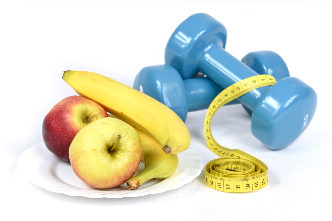 La imagen muestra un plato con dos manzanas y dos plátanos. Al lado hay una cinta métrica y dos pesas.