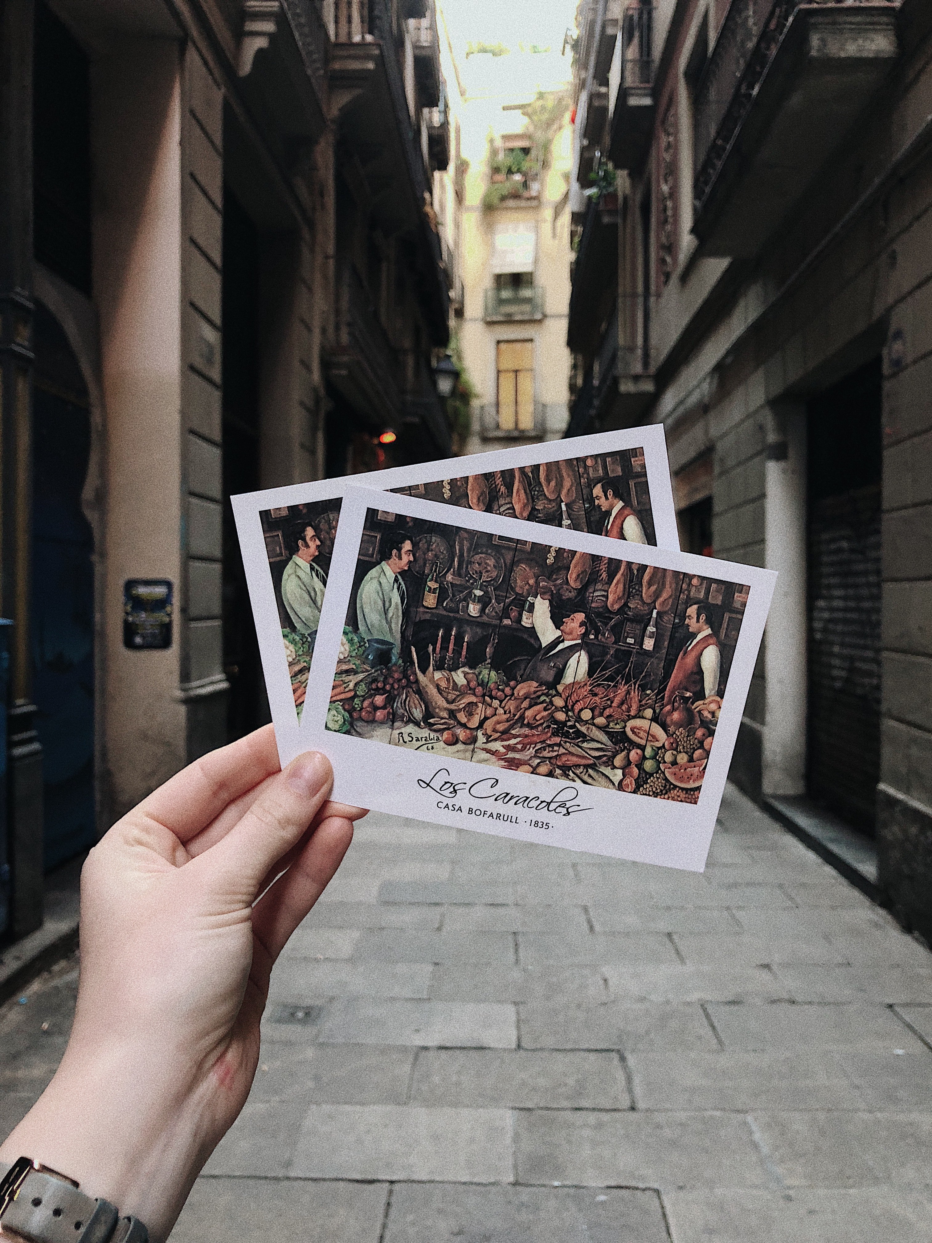 Mano sosteniendo dos postales en una calle de una ciudad.