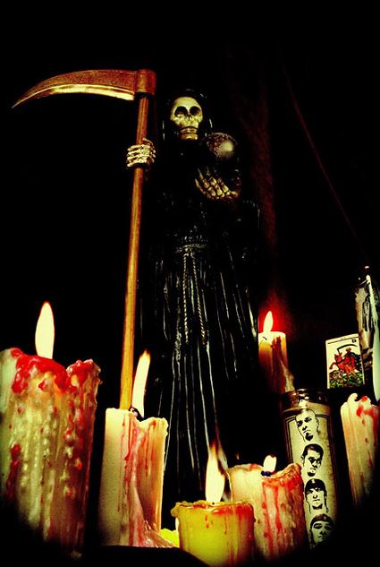 Esta imagen muestra un esqueleto con una guadaña en la mano y a sus pies velas.