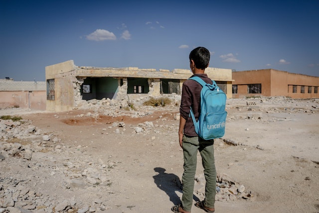 La imagen muestra a un chico con una mochila mirando su colegio en ruinas.
