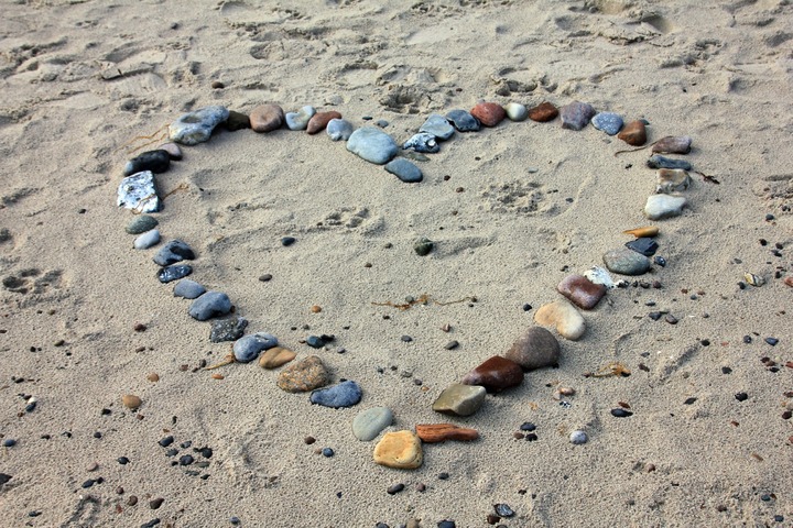 La imagen muestra un corazón sobre la arena de la playa hecho con piedrecitas.