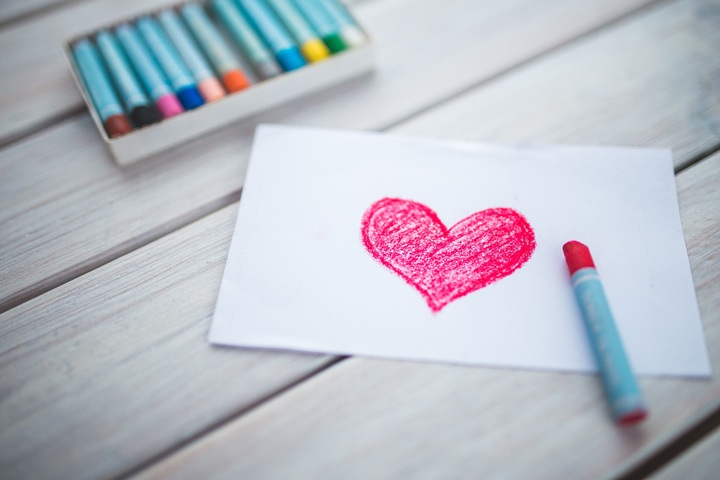 La imagen muestra un corazón rojo pintado en un papel junto a una caja de ceras de colores.