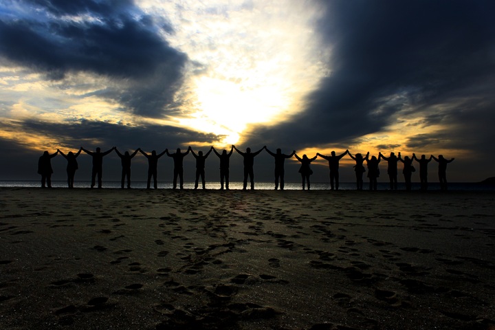 La imagen muestra un grupo de 18 personas con los brazos en alto, unidos por sus manos, en la orilla de una playa mirando hacia el mar en el momento del ocaso del día.