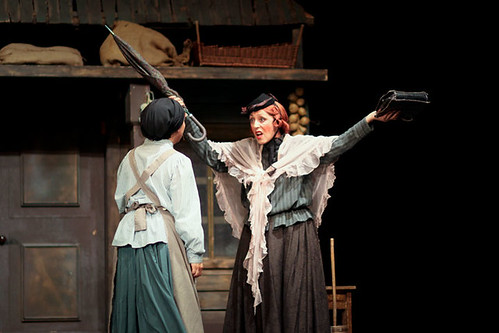 La imagen muestra una representación teatral en la que una de las actrices alza ostensiblemente los brazos.