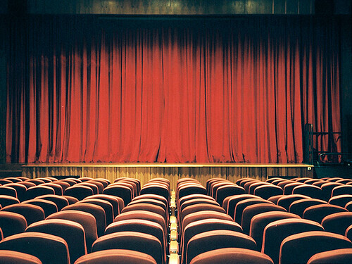 La imagen muestra una sala de teatro con el escenario tapado por unas cortinas.