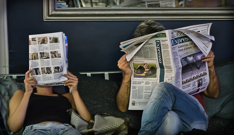 Dos personas sentadas leen respectivamente una revista y un periódico.