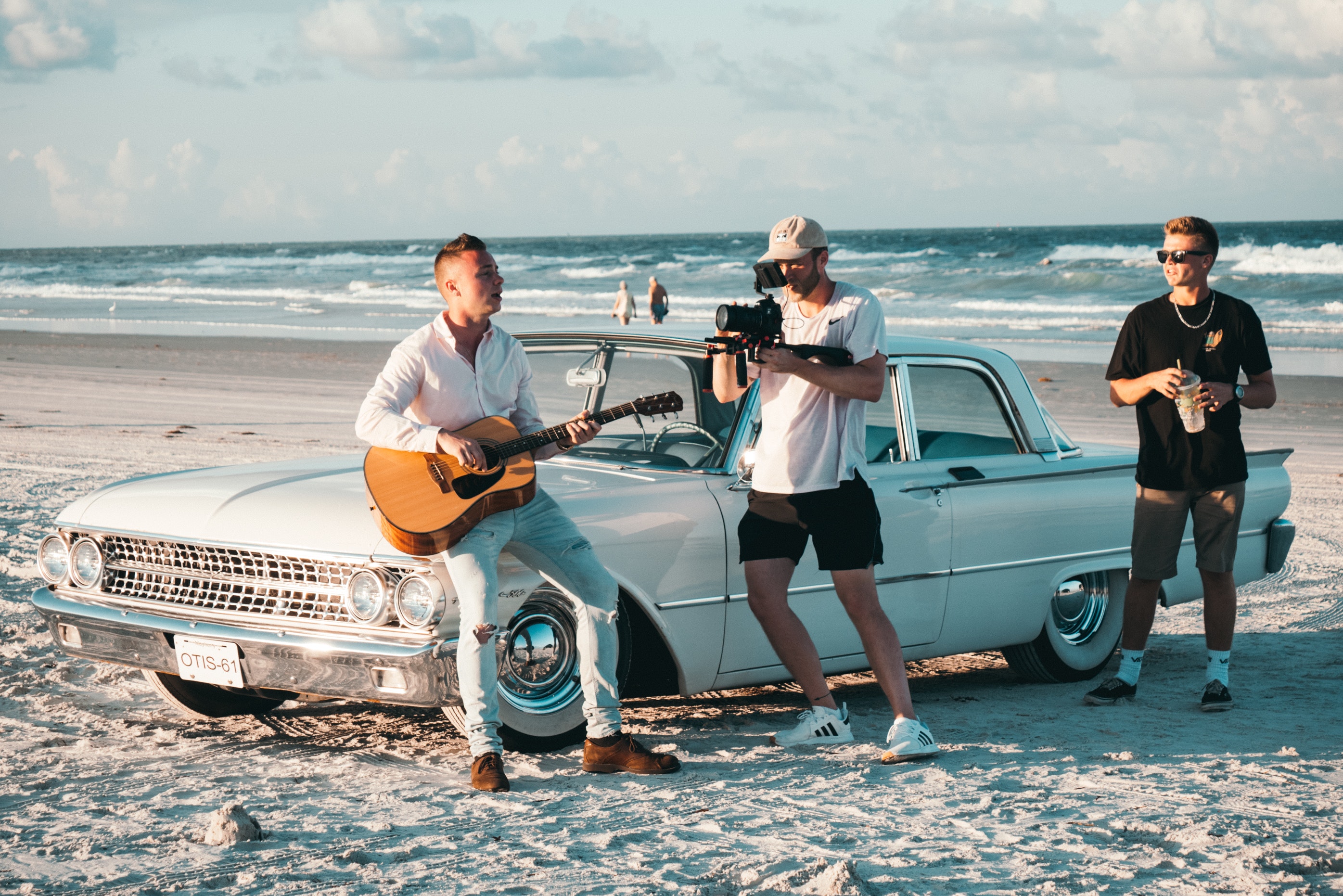 La imagen muestra a un hombre tocando la guitarra apoyado en un coche y a otro hombre grabando la escena.