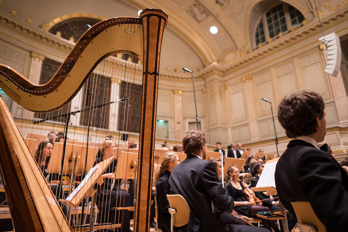 La imagen muestra un arpa en primer plano y de fondo una orquesta en un auditorio