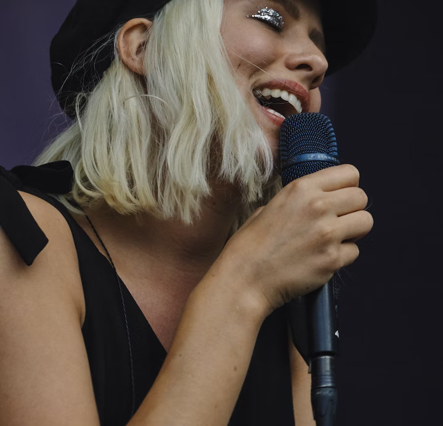 La imagen muestra a una chica joven con gorro negro, rubia de pelo corto de perfil cantando y sujetando un micrófono