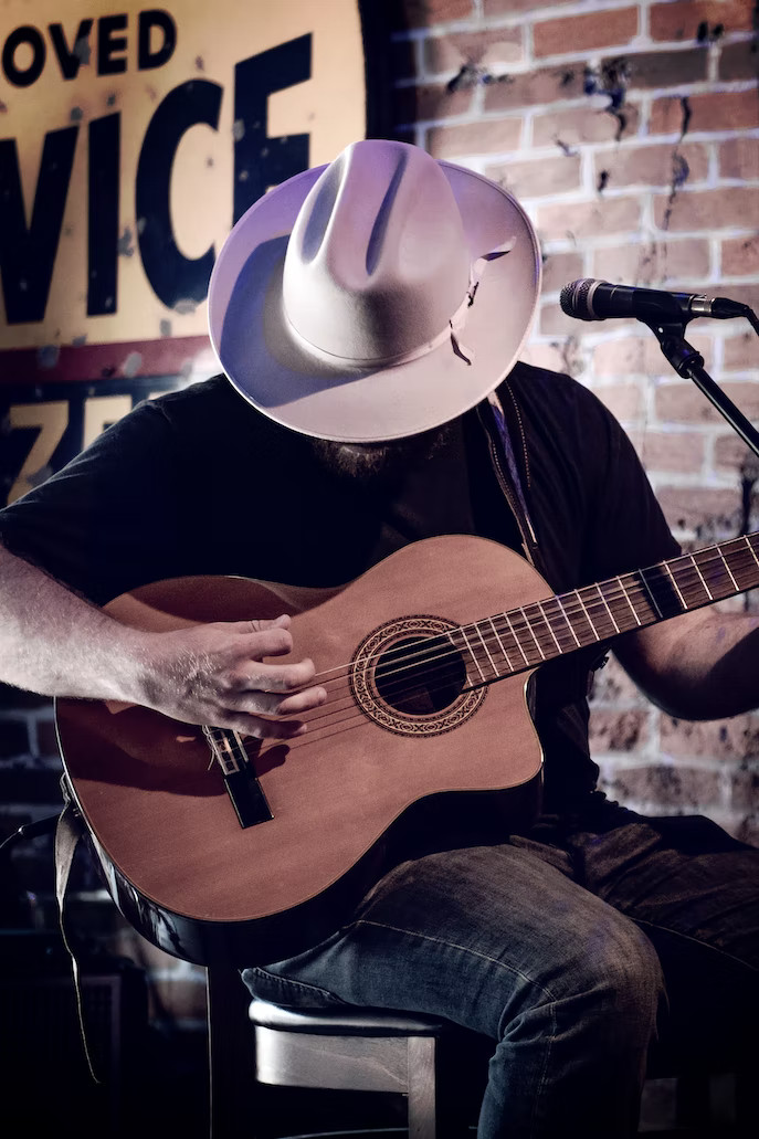 La imagen muestra hombre con la cabeza agachada con un gorro country y tocando una guitarra