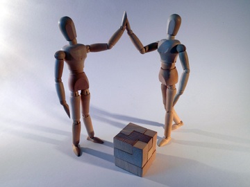 La imagen muestra dos figuras de madera chocando la mano, delante de ellas un bloque de madera hecho de varias piezas.