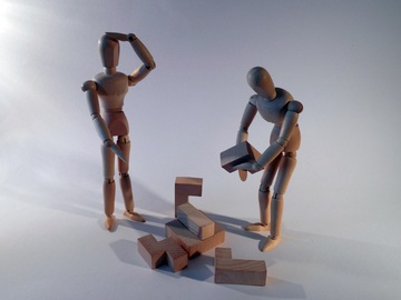 La imagen muestra dos figuras humanas de madera enfrentándose a la construcción de un bloque de madera compuesto de varias piezas que encajan unas con otras.