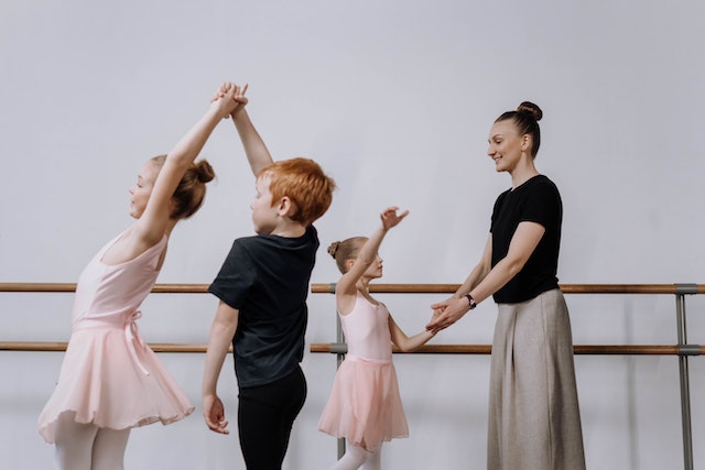 La imagen muestra a una alumna en clase de baile ayudando a alumnos más jóvenes. 