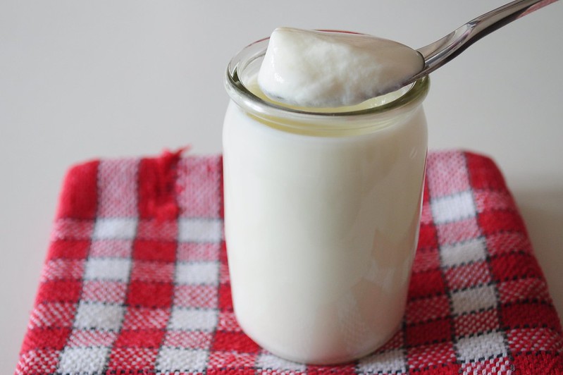 En la imagen se puede ver un yogur natural en un tarro transparente sobre un mantel de cuadros rojos y blancos