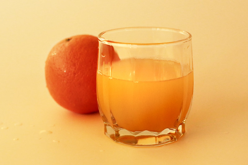 En la imagen se puede ver un vaso de zumo de naranja con narajas detrás