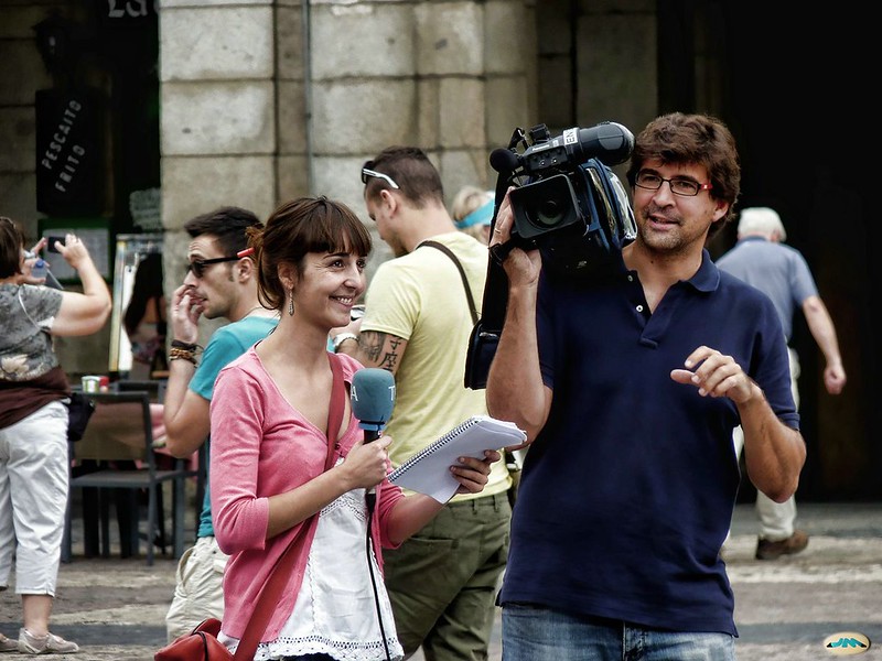 Reportera sonríe mientras lleva documentos en la mano. Su compañero lleva la cámara al hombro.