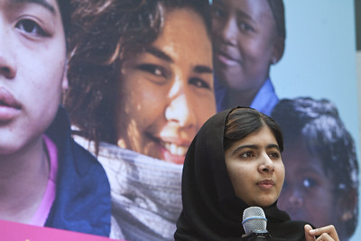 Imagen del rostro de Malala, una niña de origen pakistaní, que tapa su cabeza con un pañuelo negro. Lleva un micrófono en la mano. Tras ella se ve una imagen enorme con rostros de distintas niñas.