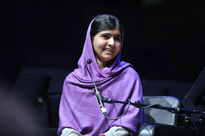 Imagen de Malala, una chica de origen pakistaní vestida de color morado y con un pañuelo tapando la cabeza, el pelo que se le puede ver es de color negro. Está sentada freten a un micrófono. sonríe.