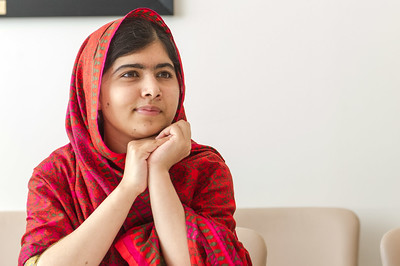 Imagen de Malala, una niña de origen pakistaní, sentada en un sofá. Lleva el pelo y los hombros cubiertos por un pañuelo rojo. Las manos las tiene a la altura de la barbilla y se apoya sobre ellas.
