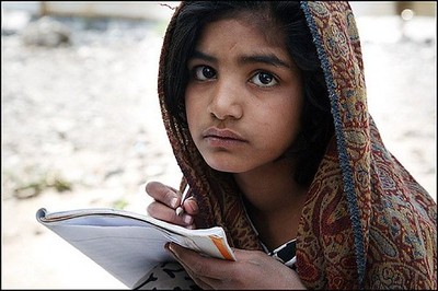 Imagen del rostro de una niña que lleva el cabello cubierto por un pañuelo. Está sentada y tiene un cuaderno y un lápiz en la mano.
