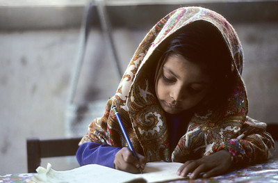 Imagen de una niña escribiendo sobre un cuaderno con un lápiz. Cubre su pelo y hombros con un pañuelo.