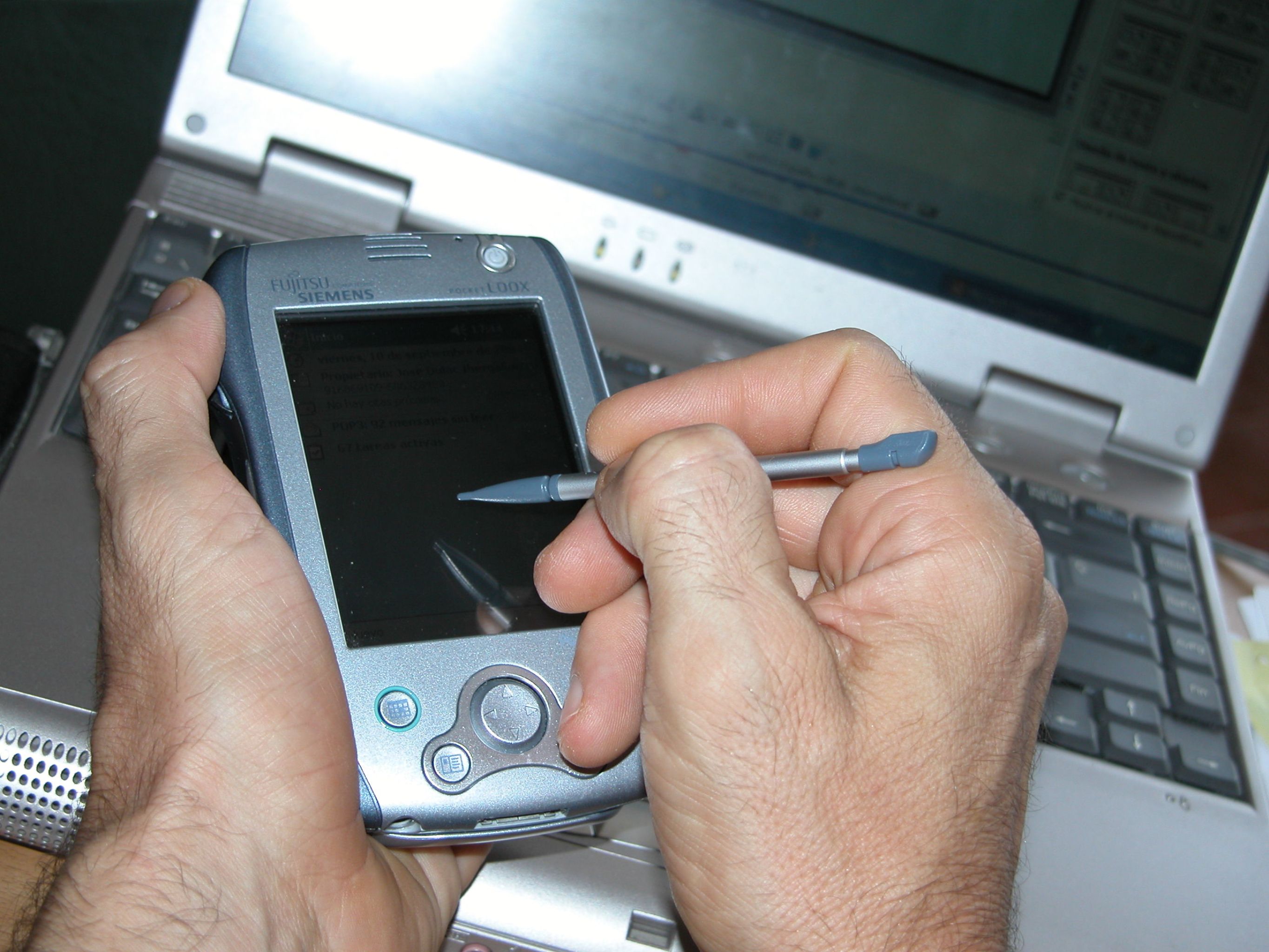 Alguien sostiene una PDA y escribe sobre ella con un lápiz digital. En el fondo aparece un portátil.