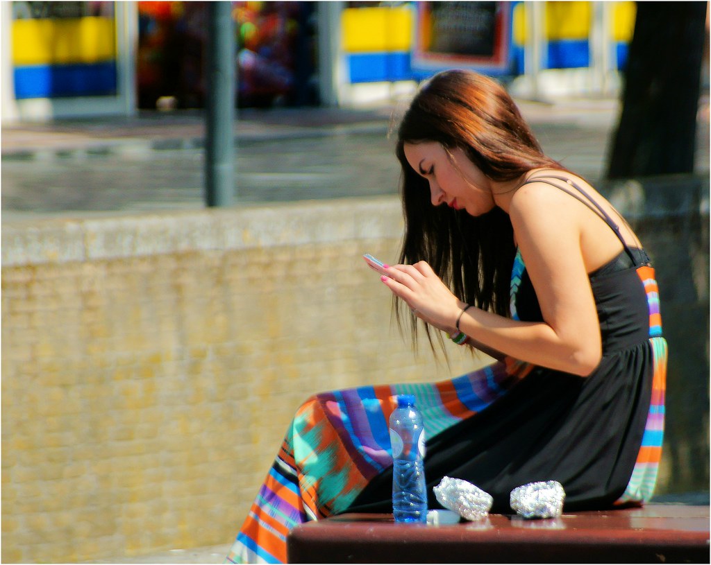 Chica sentada en un banco en la calle, tiene el pelo largo y castaño, y está con la cabeza inclinada hacia un teléfono móvil que está manipulando.
