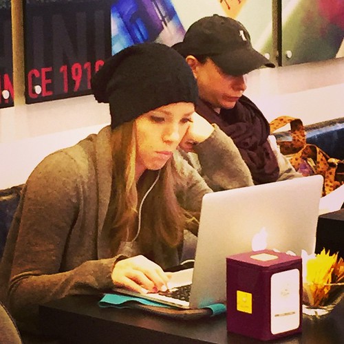 Fotografía de una chica de pelo largo y rubio, con un gorro de invierno negro, sentada en una cafetería usando un ordenador portátil. Lleva puestos unos auriculares. Junto a ella se ve un chico sentado con una gorra.