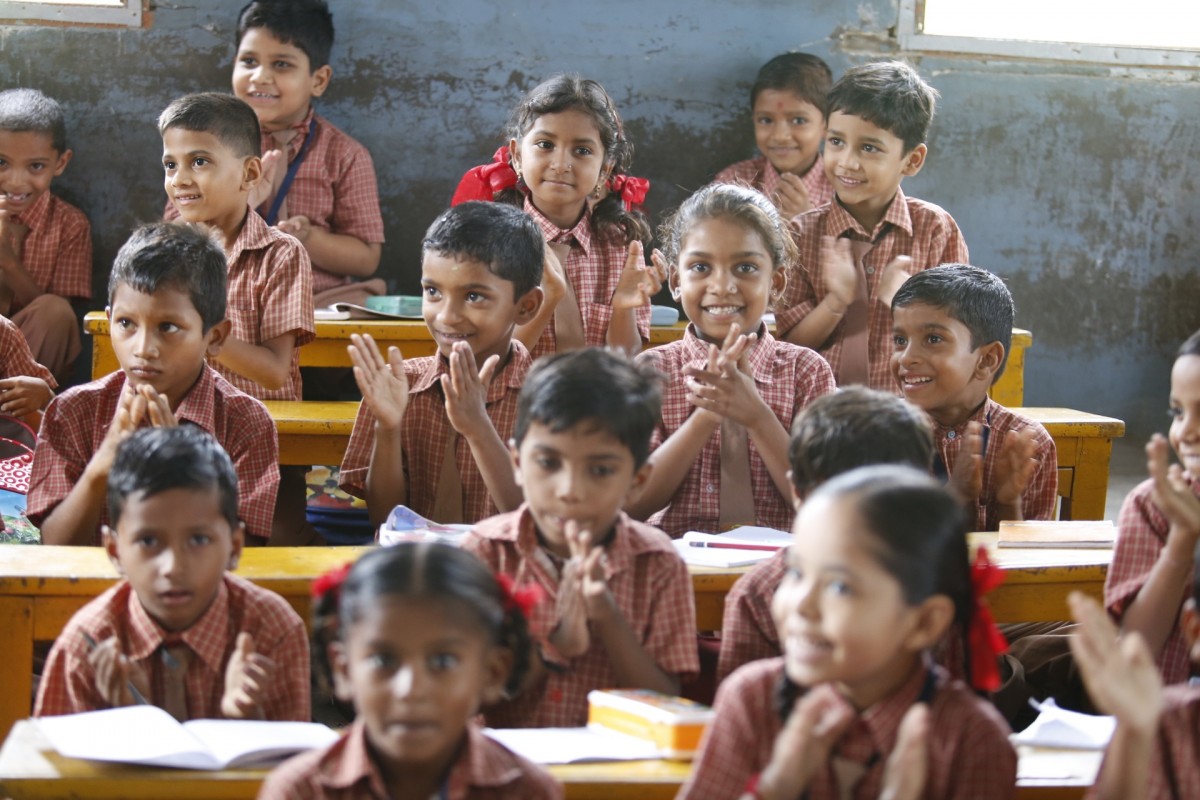 Imagen de un aula, en la que hay varios niños y niñas sentados en sus mesas sonriendo.