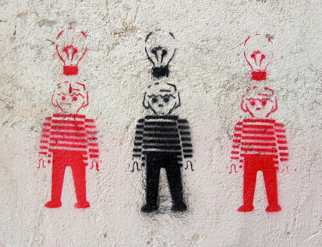 Dibujo de tres muñecos de playmobil, dos rojos en los laterales y uno negro en el centro. Cada uno tiene una bombilla de su color sobre su cabeza.