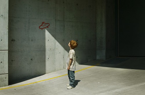 Dibujo de un niño en una calle observando un dibujo en la pared. El dibujo de la pared es el de un platillo volante.