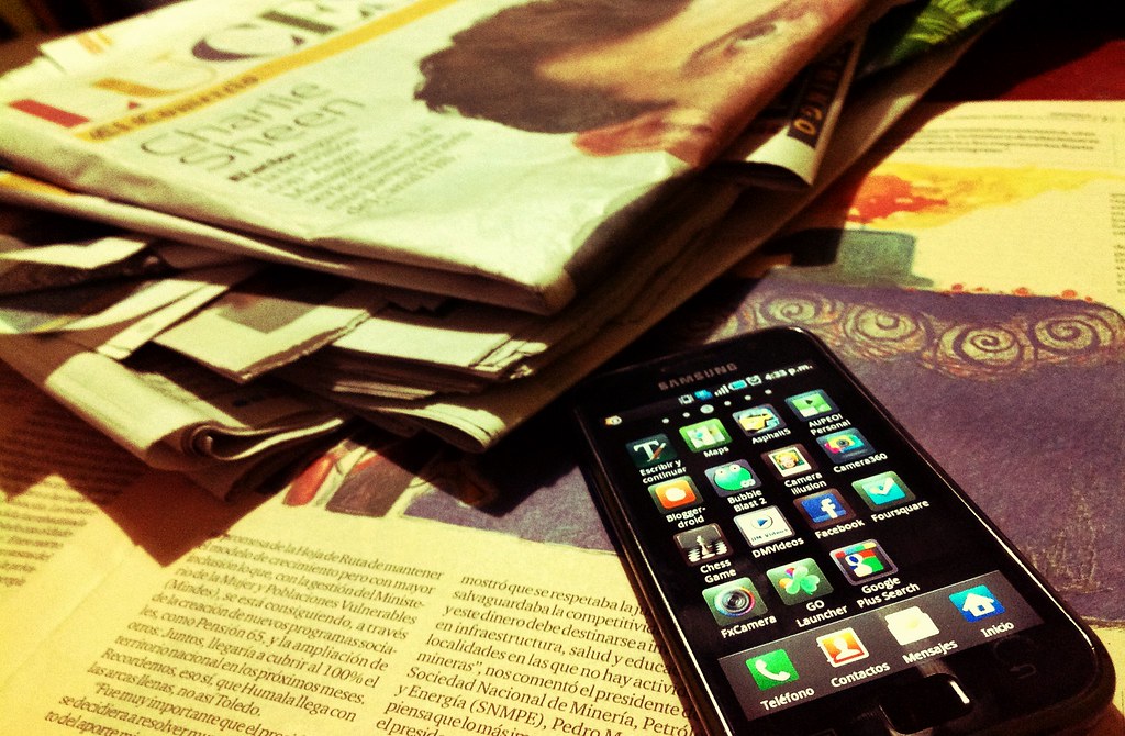 Un teléfono móvil con la pantalla encendida, se ve en ella los iconos de diversas aplicaciones sobre un fondo negro. El móvil está sobre unos periódicos.