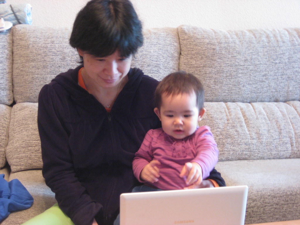 Fotografía de una mujer de pelo corto y rasgos asiáticos, sentada en un sofá y con un bebé vestido de rosa en su regazo, mirando la pantalla de un ordenador.