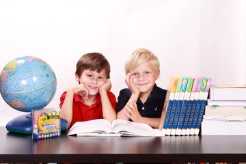 Dos niños, mirando a cámara, en una mesa con diverso material escolar: globo terráqueo, caja de colores y varios libros.