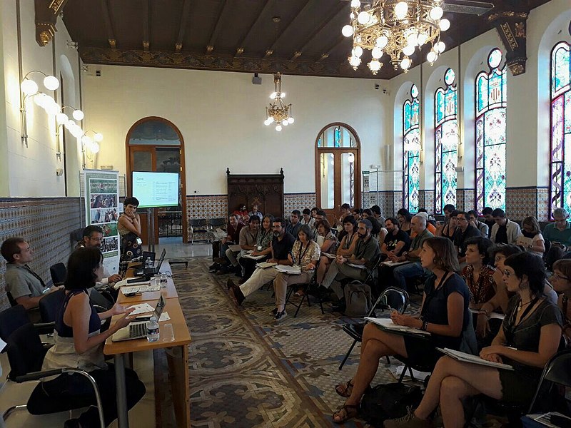 La imagen muestra un conjunto de personas atentas a otro grupo reducido que se encuentra frente a una mesa alargada. 