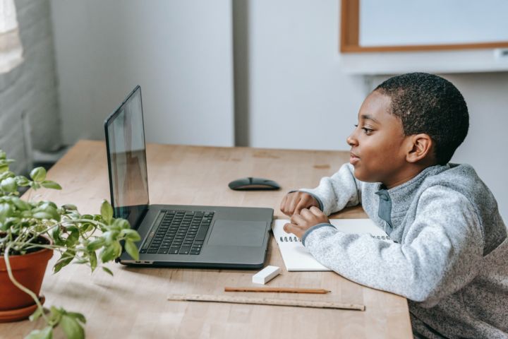 En la fotografía aparece un niño de piel oscura mirando un video y tomando apuntes de lo que aprende a través del ordenador.