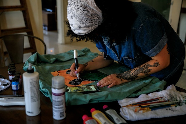 Aparece una mujer con un pañuelo en la cabeza y esta pintando una camiseta con pintura, realiza un dibujo sobre la camiseta.