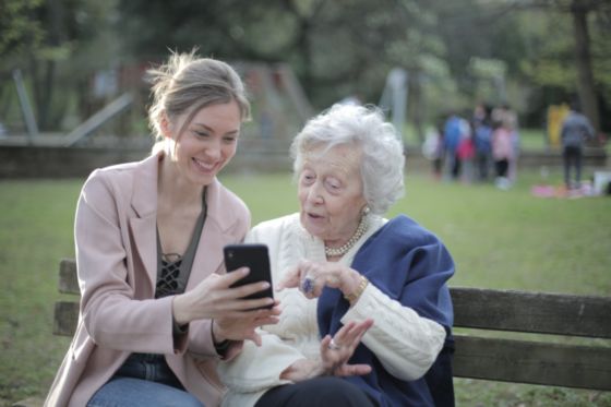 En la imagen aparece una mujer joven acompañada de una anciana, la mujer le enseña una imagen del móvil sentadas en un parque. Sus caras expresan que el contenido tiene relación con ambas.