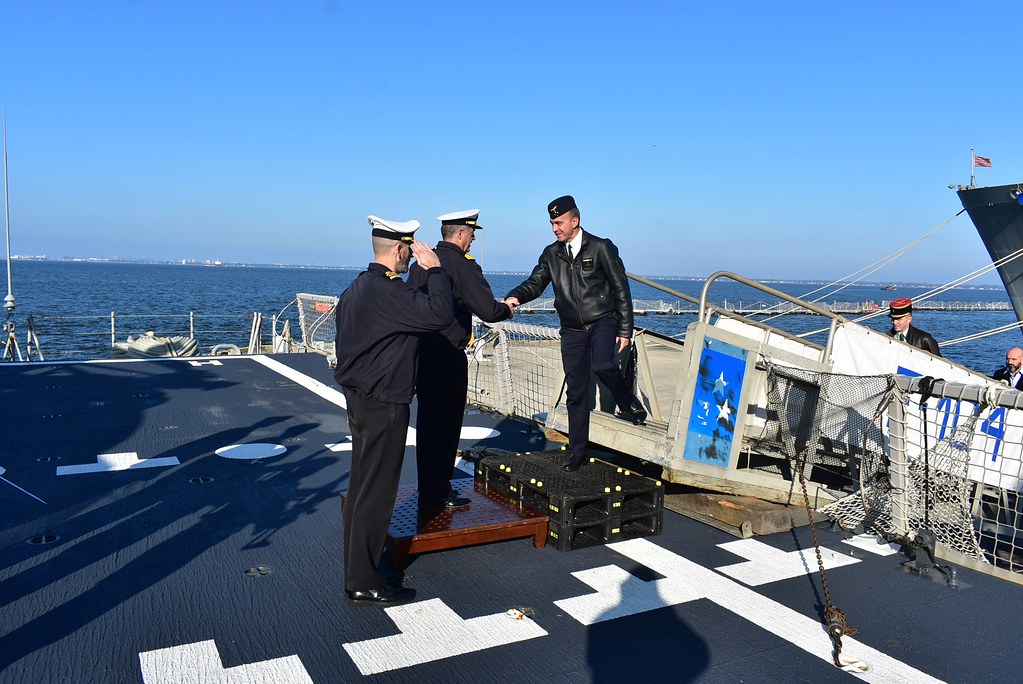 La imagen muestra a dos comandantes que reciben a otro en un portaviones.