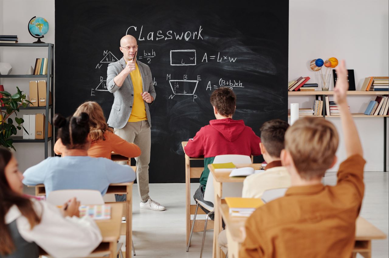 La imagen muestra a un maestro haciendo una pregunta en clase y a un alumno levantando la mano .