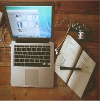 La imagen muestra un ordenador portátil abierto, una libreta abierta con un lápiz encima y unos cascos con cable.
