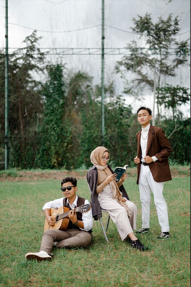 La imagen muestra a tres adolescentes sentados sobre el césped de un parque. Uno toca la guitarra, otra lee un libro y el tercero mira al horizonte.