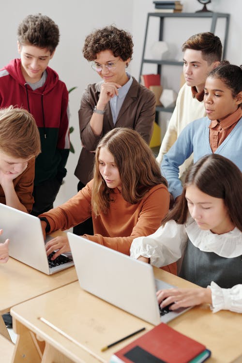 La imagen muestra a un grupo de adolescentes mirando pantallas de ordenador. Algunos están sentados y otros de pie.