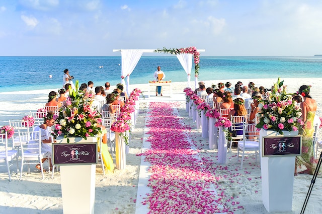 Paisaje de playa, con decoración de boda. Tiene sillas en filas, una alfombra roja, y flores por el suelo. Todas las personas están esperando a los novios porque es una boda en la playa.