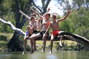 La imagen muestra a unos adolescentes en bañador subidos a un árbol pasándolo bien en un río. 