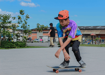 La imagen muestra a un niño con gorra patinando en su monopatín en un parque. Al fondo se ve otro niño saltando con su monopatín, unos edificios bajos, árboles y un parque infantil con columpios.